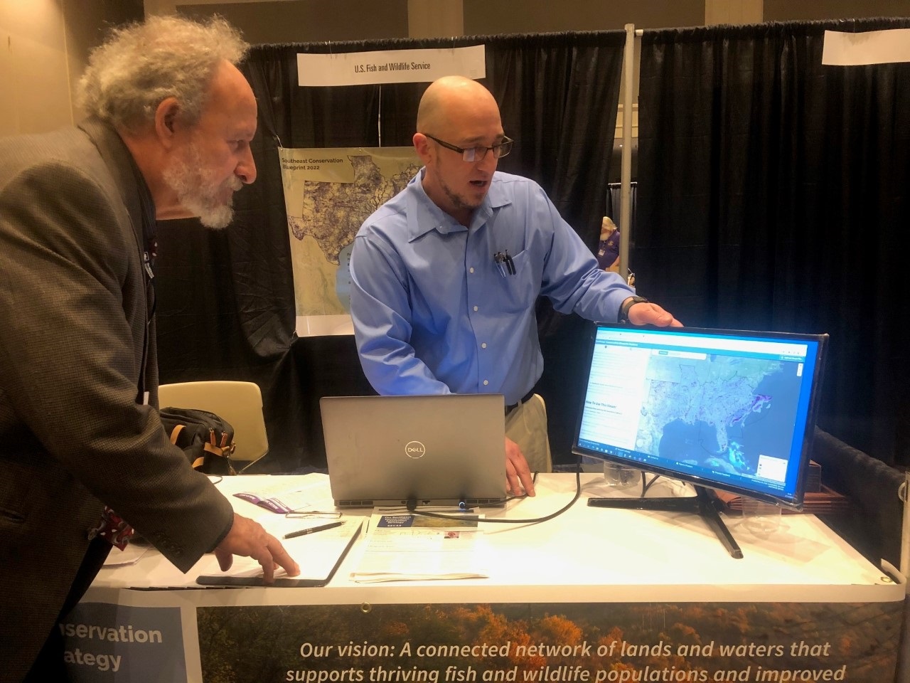 Photo of Adam at an exhibit booth showing a meeting attendee the Southeast Conservation Blueprint on a large screen.